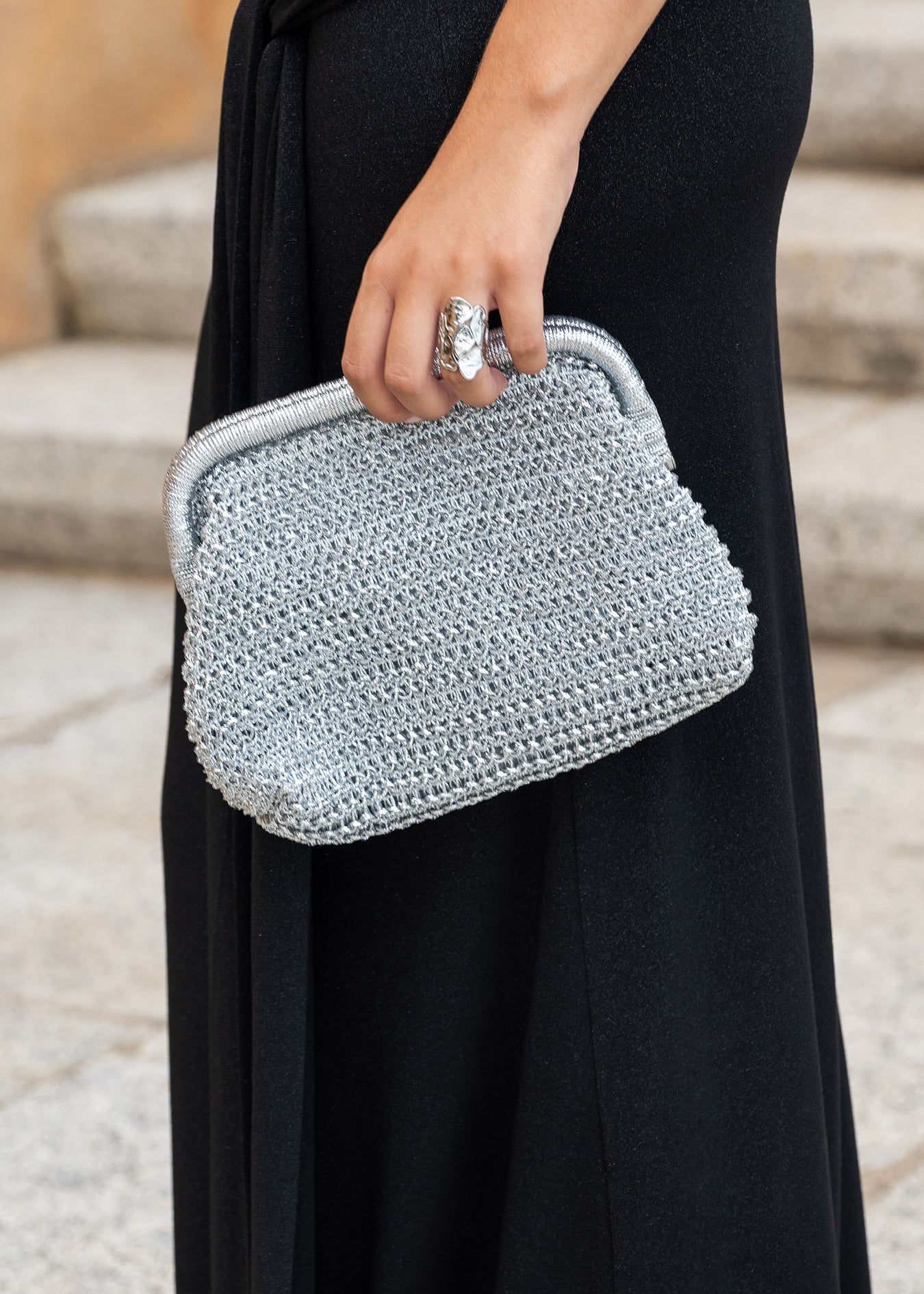 Primer plano del bolso Mikonos plata tejido con asa metálica y detalle del anillo plateado en la mano de la modelo, con un fondo de escaleras de piedra.