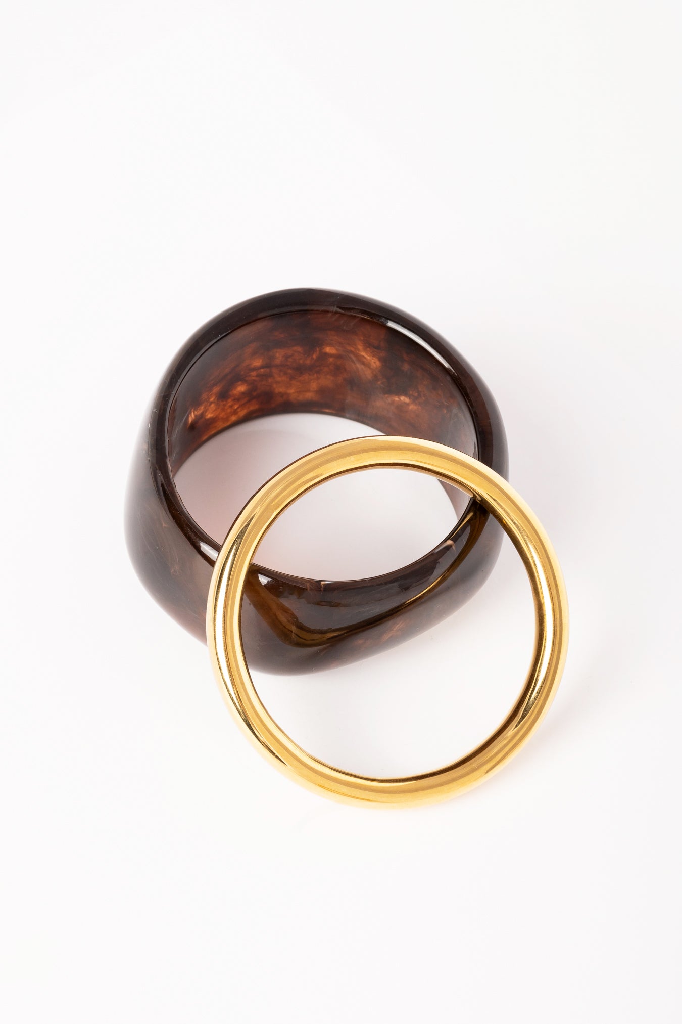 “Composición de dos brazaletes: Brazalete Lola en resina marrón oscuro con efecto marmoleado y brazalete Bimba dorado, dispuestos sobre fondo blanco.”