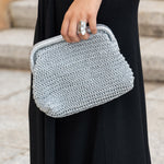  Primer plano del bolso Mikonos plata tejido con asa metálica y detalle del anillo plateado en la mano de la modelo, con un fondo de escaleras de piedra.