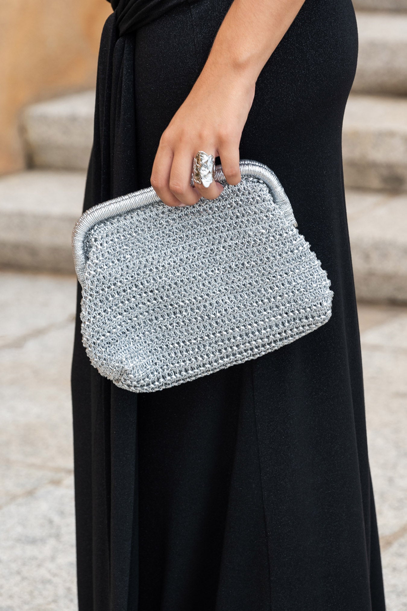  Primer plano del bolso Mikonos plata tejido con asa metálica y detalle del anillo plateado en la mano de la modelo, con un fondo de escaleras de piedra.