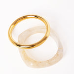 “Brazalete dorado apilado sobre otro brazalete de resina translúcida con textura marmoleada, ambos sobre fondo blanco.”