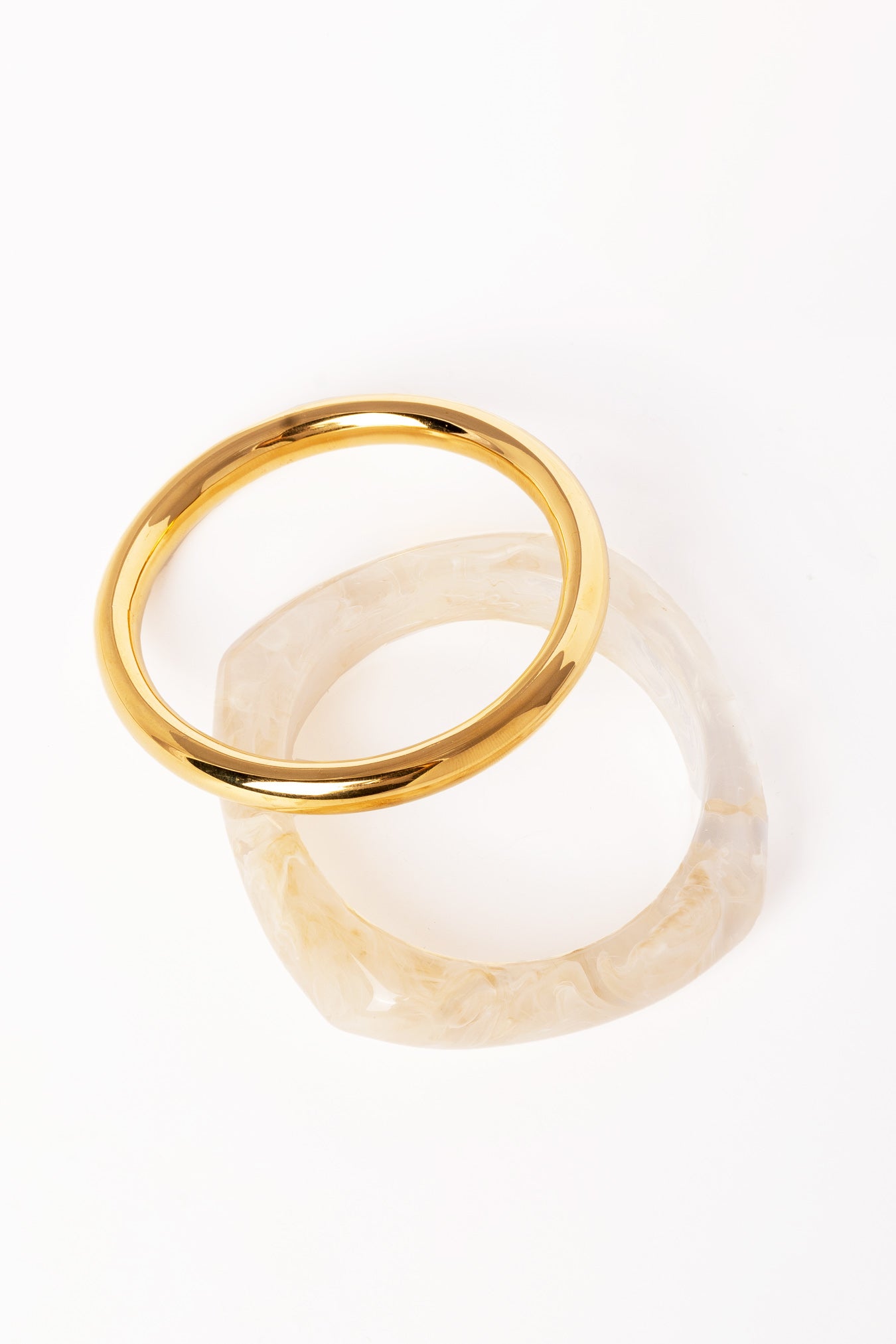 “Brazalete dorado apilado sobre otro brazalete de resina translúcida con textura marmoleada, ambos sobre fondo blanco.”