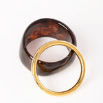  “Composición de dos brazaletes: Brazalete Lola en resina marrón oscuro con efecto marmoleado y brazalete Bimba dorado, dispuestos sobre fondo blanco.”