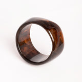 “Brazalete Lola de resina en tonos marrón oscuro, en posición inclinada sobre fondo blanco para resaltar su forma redondeada.”