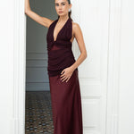 Modelo con Vestido largo en satén color burdeos, con escote halter cruzado y abertura triangular en el busto. El escote y el fruncido de la cadera están delicadamente trabajados en tul. La falda recta estiliza y aporta elegancia y movimiento. Ideal para bodas, galas y eventos de noche.
