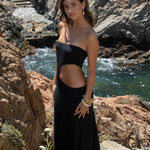 “Mujer de pie sobre un sendero de piedra, llevando un vestido largo negro con abertura lateral y accesorios dorados, con el mar y las rocas detrás.”

