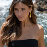 “Mujer llevando pendientes Kalma Oro junto al mar, con el cabello suelto y vestido palabra de honor negro.”