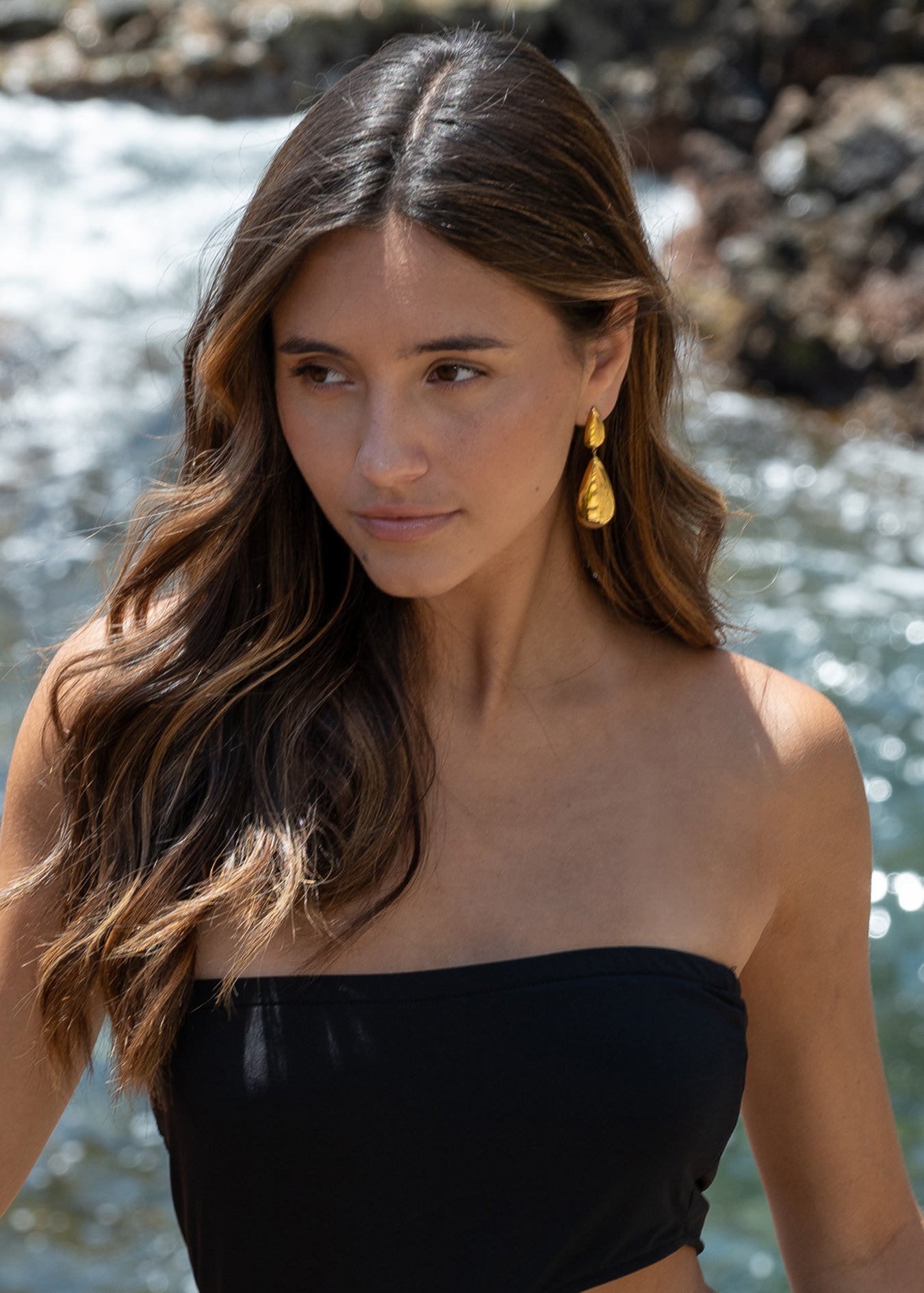 “Mujer llevando pendientes Kalma Oro junto al mar, con el cabello suelto y vestido palabra de honor negro.”