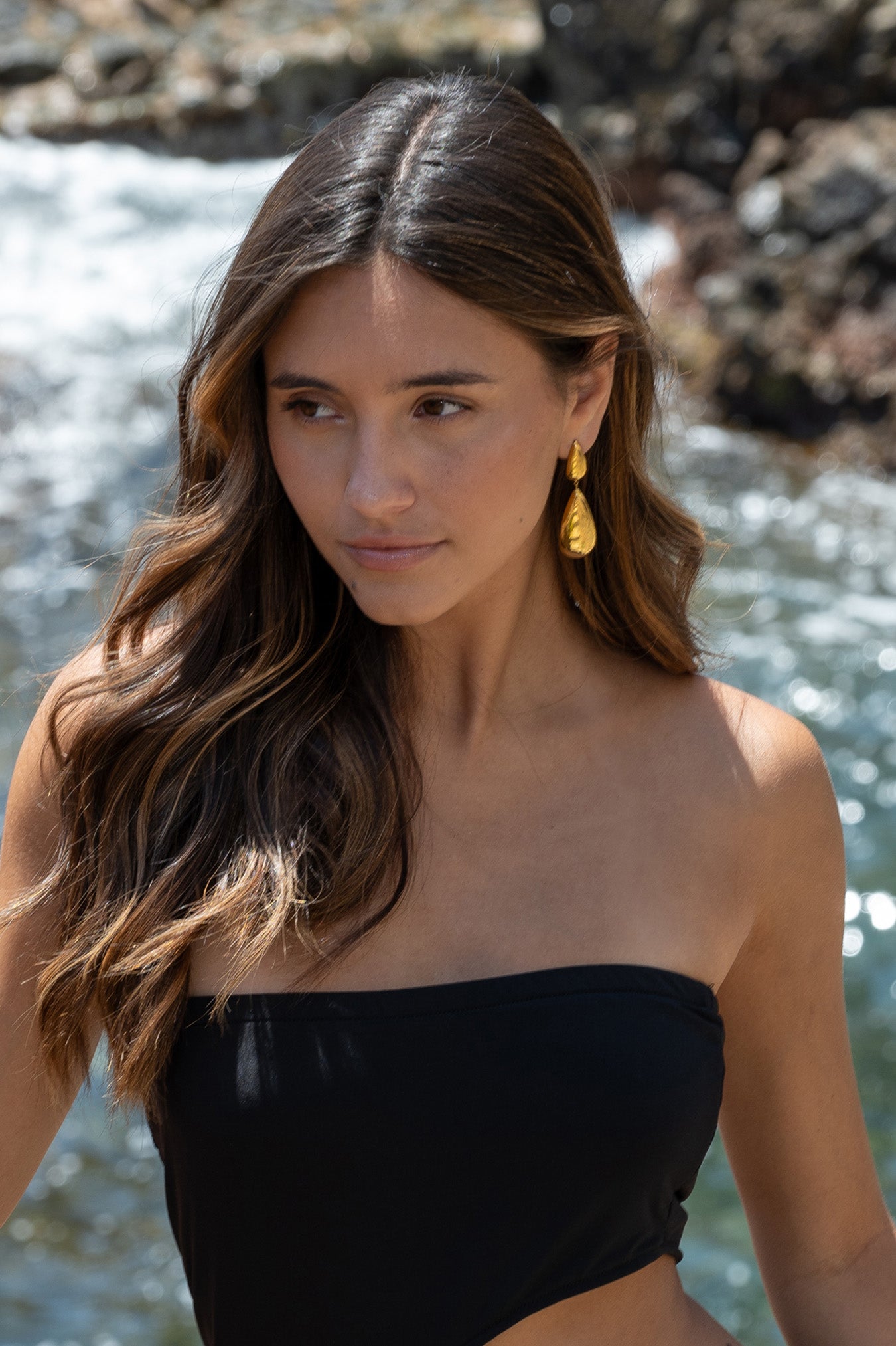 “Mujer llevando pendientes Kalma Oro junto al mar, con el cabello suelto y vestido palabra de honor negro.”