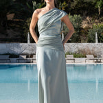 Vestido largo de invitada color verde salvia con escote asimétrico y sofisticado drapeado frontal frente a una piscina.