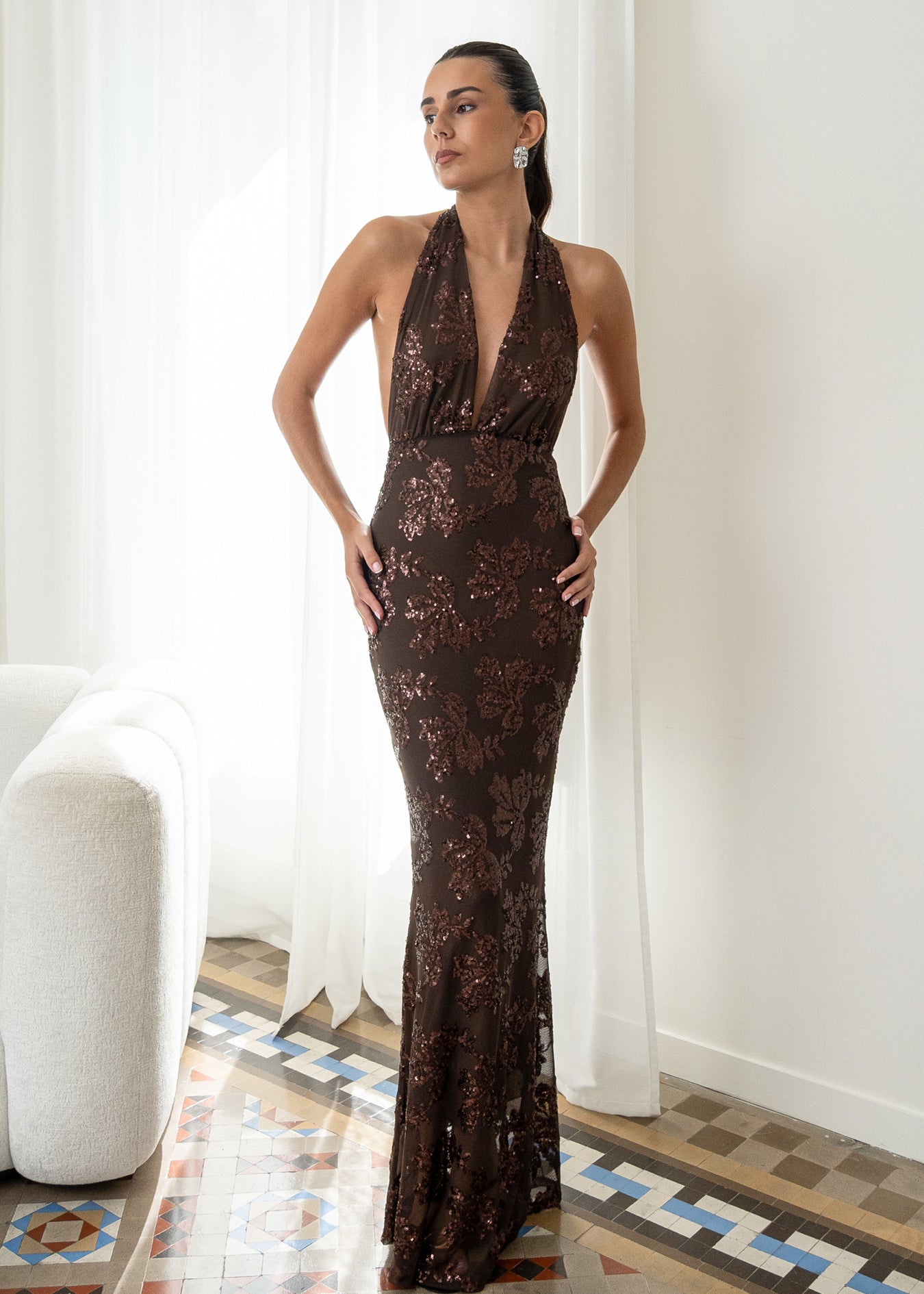 Modelo con vestido Fiori Marrón su escote halter atado al cuello y su espalda descubierta aportan un aire sofisticado y moderno. Confeccionado en gasa ligera decorada con lentejuelas.