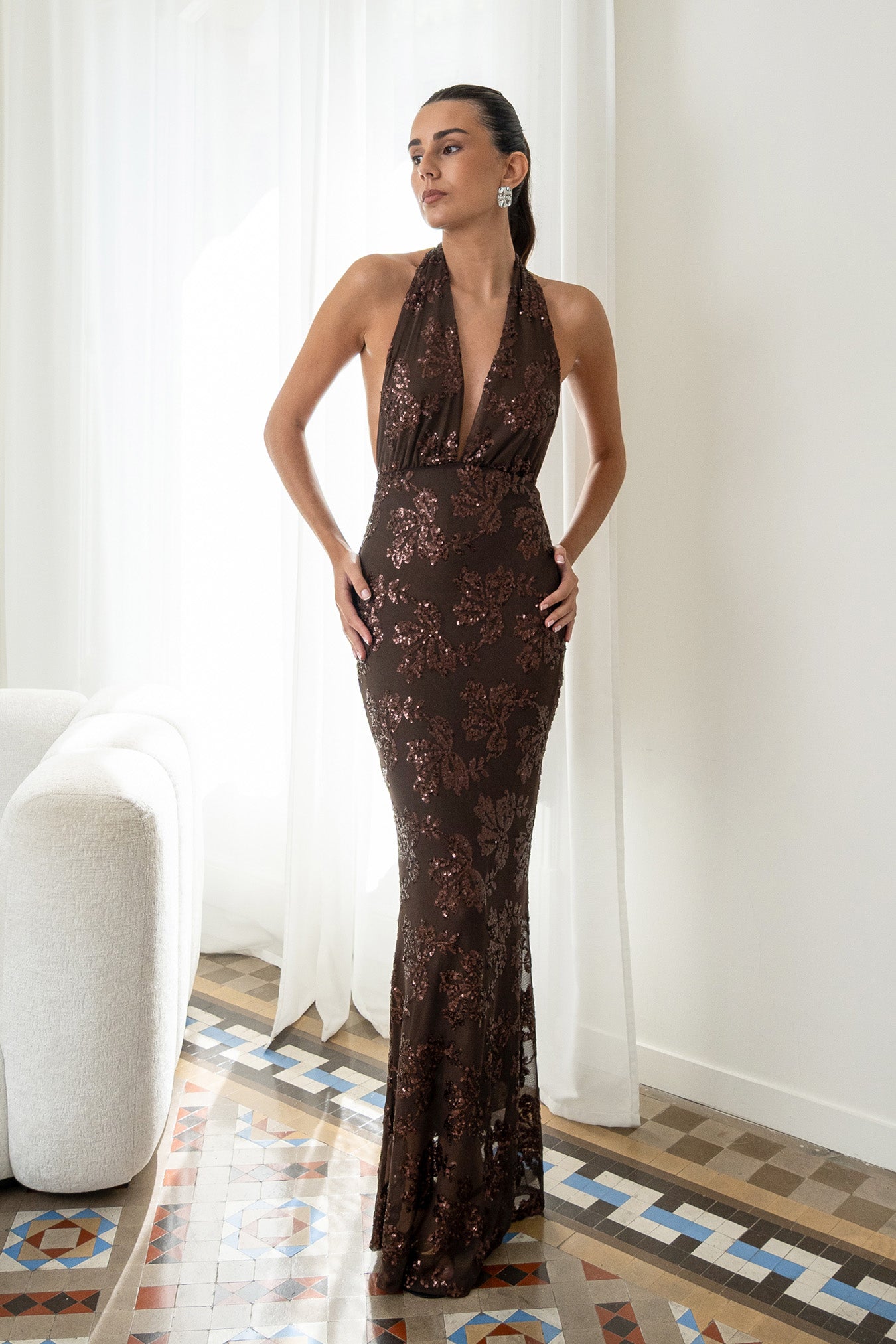 Modelo con vestido Fiori Marrón su escote halter atado al cuello y su espalda descubierta aportan un aire sofisticado y moderno. Confeccionado en gasa ligera decorada con lentejuelas.