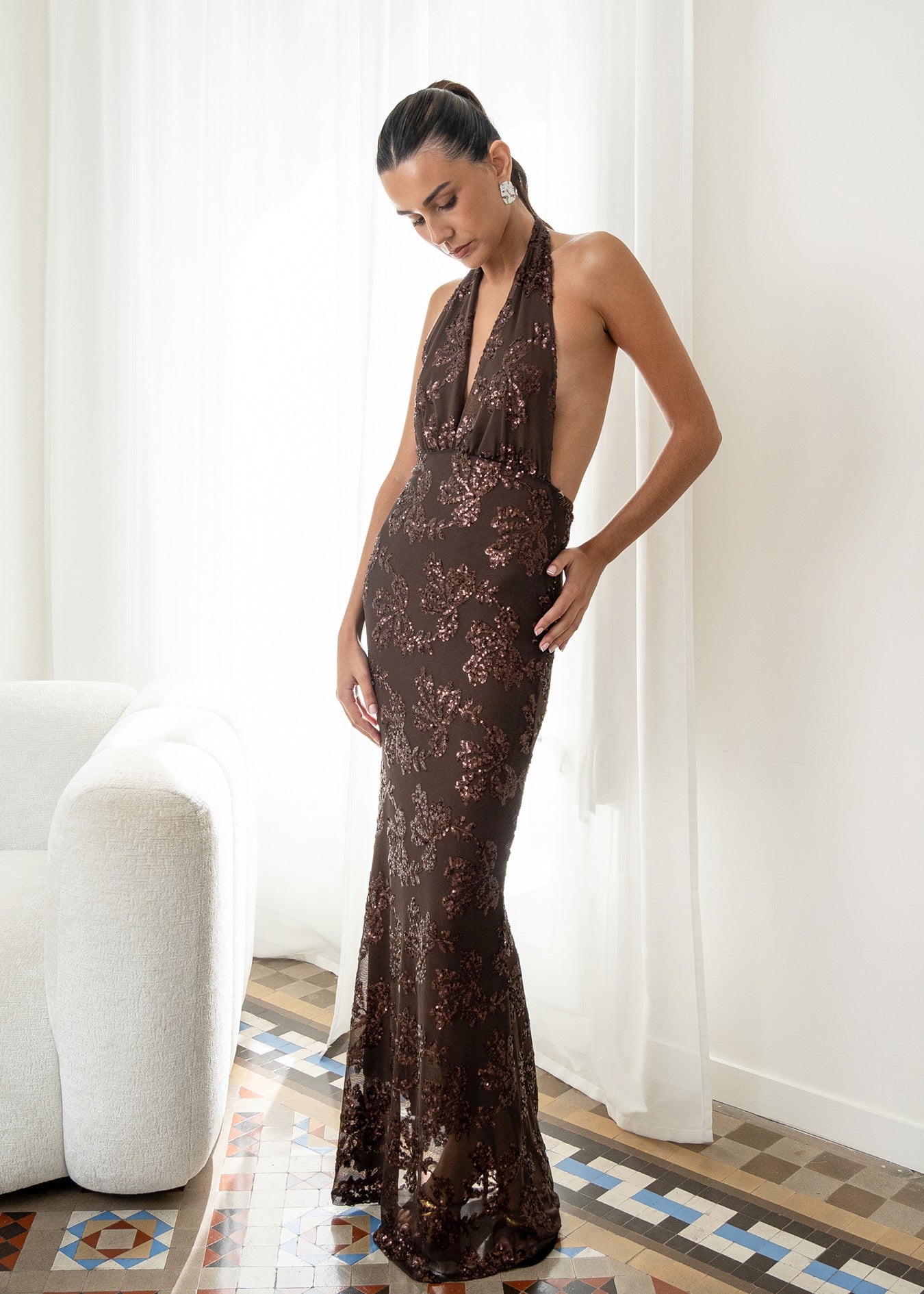 Modelo con vestido Fiori Marrón su escote halter atado al cuello y su espalda descubierta aportan un aire sofisticado y moderno. Confeccionado en gasa ligera decorada con lentejuelas.