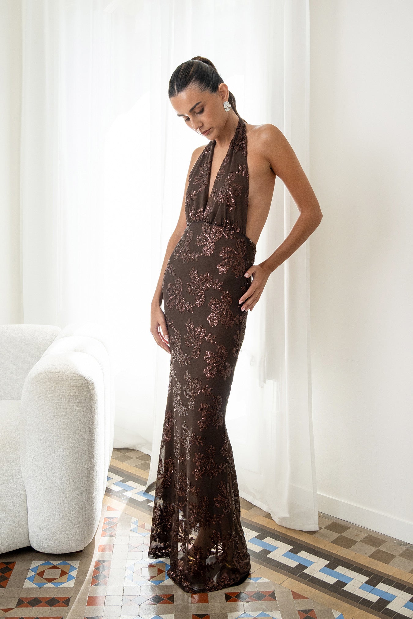 Modelo con vestido Fiori Marrón su escote halter atado al cuello y su espalda descubierta aportan un aire sofisticado y moderno. Confeccionado en gasa ligera decorada con lentejuelas.