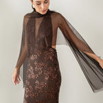 Modelo con vestido Fiori Marrón su escote halter atado al cuello y su espalda descubierta aportan un aire sofisticado y moderno. Confeccionado en gasa ligera decorada con lentejuelas.