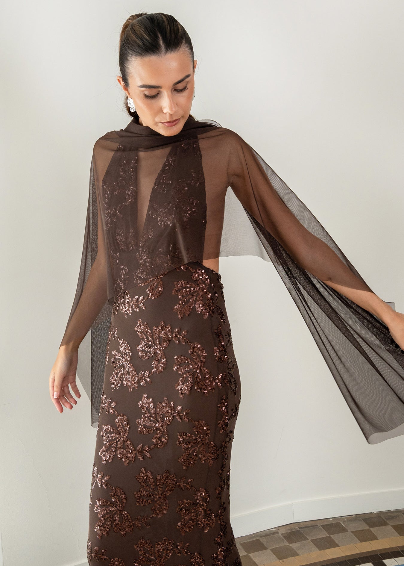 Modelo con vestido Fiori Marrón su escote halter atado al cuello y su espalda descubierta aportan un aire sofisticado y moderno. Confeccionado en gasa ligera decorada con lentejuelas.