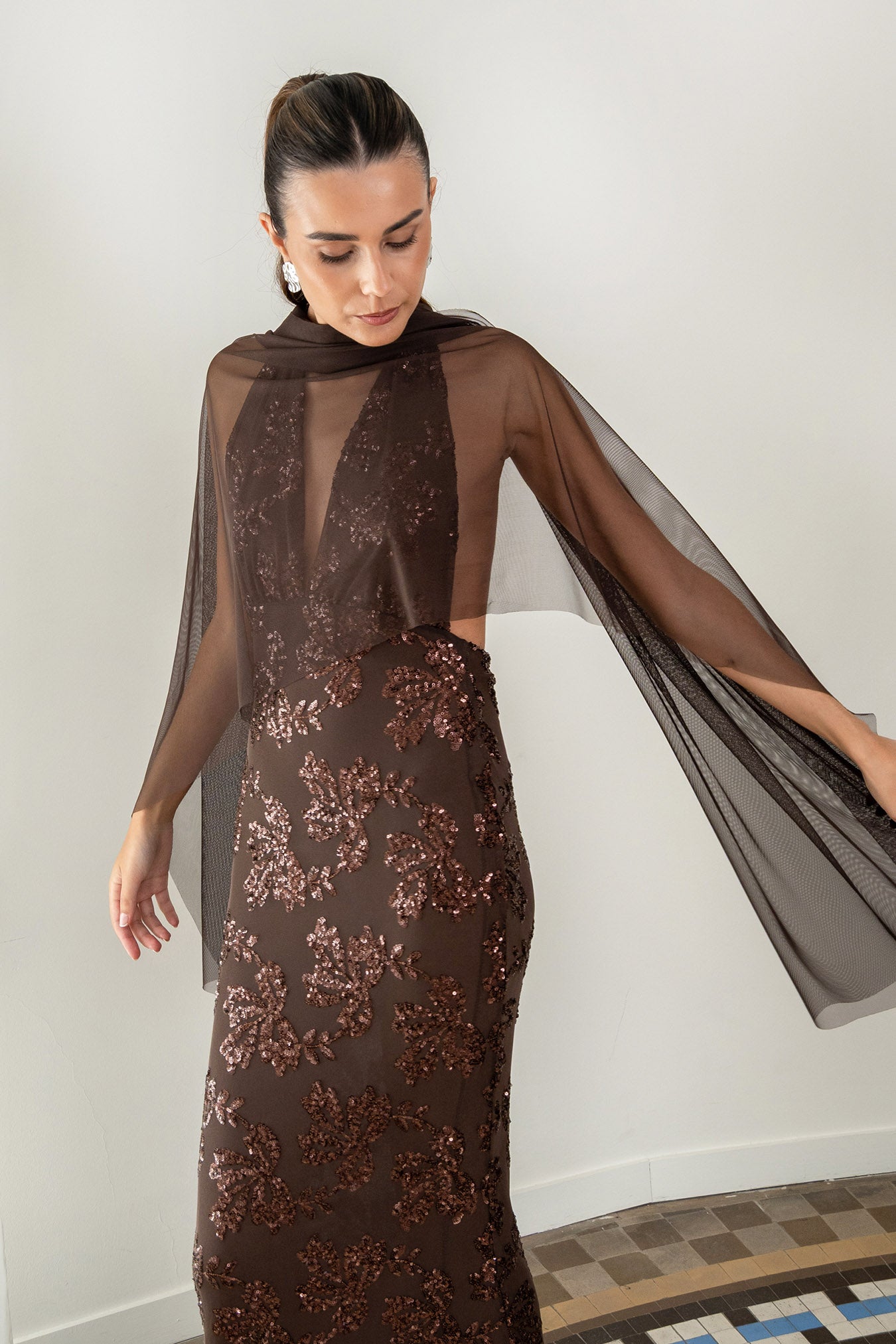 Modelo con vestido Fiori Marrón su escote halter atado al cuello y su espalda descubierta aportan un aire sofisticado y moderno. Confeccionado en gasa ligera decorada con lentejuelas.
