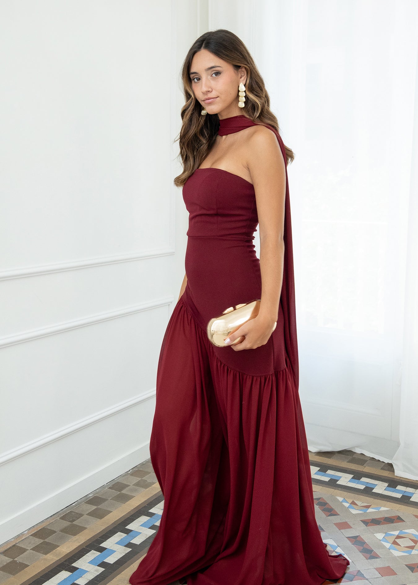 Modelo con vestido largo burdeos,  ideal para invitadas a bodas, fiestas de noche o eventos de gala. Escote palabra de honor que realza los hombros y el cuello, acompañado de un elegante fular integrado, aportando un toque sofisticado y original.