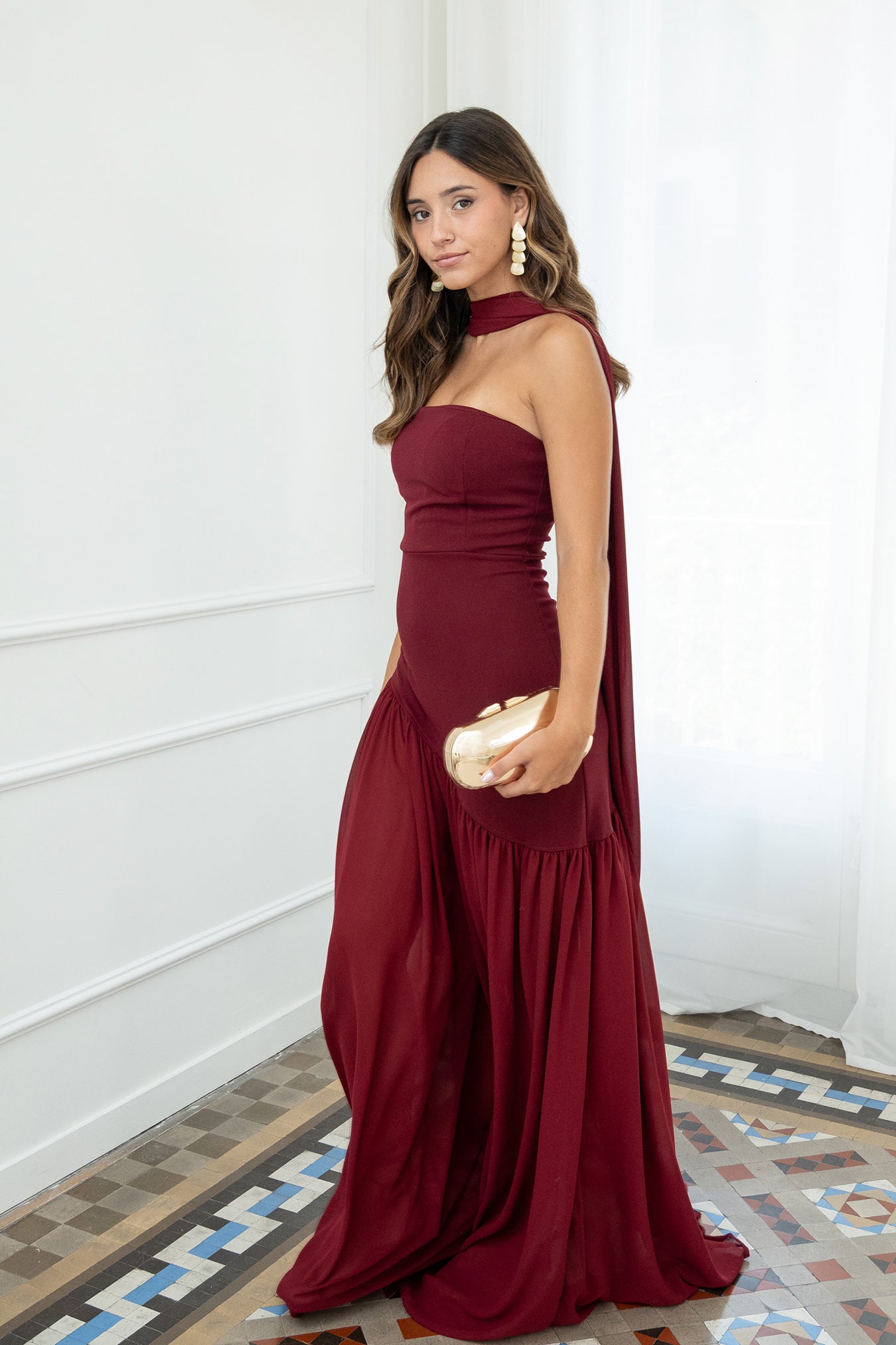 Modelo con vestido largo burdeos,  ideal para invitadas a bodas, fiestas de noche o eventos de gala. Escote palabra de honor que realza los hombros y el cuello, acompañado de un elegante fular integrado, aportando un toque sofisticado y original.