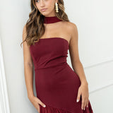 Modelo con vestido largo burdeos,  ideal para invitadas a bodas, fiestas de noche o eventos de gala. Escote palabra de honor que realza los hombros y el cuello, acompañado de un elegante fular integrado, aportando un toque sofisticado y original.
Confeccionado en un tejido fluido y ligero, el vestido presenta una falda amplia con vuelo y semitransparente  que añade movimiento y sensualidad al caminar.