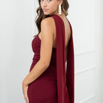 Modelo con vestido largo burdeos,  ideal para invitadas a bodas, fiestas de noche o eventos de gala. Escote palabra de honor que realza los hombros y el cuello, acompañado de un elegante fular integrado, aportando un toque sofisticado y original.