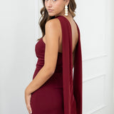 Modelo con vestido largo burdeos,  ideal para invitadas a bodas, fiestas de noche o eventos de gala. Escote palabra de honor que realza los hombros y el cuello, acompañado de un elegante fular integrado, aportando un toque sofisticado y original.