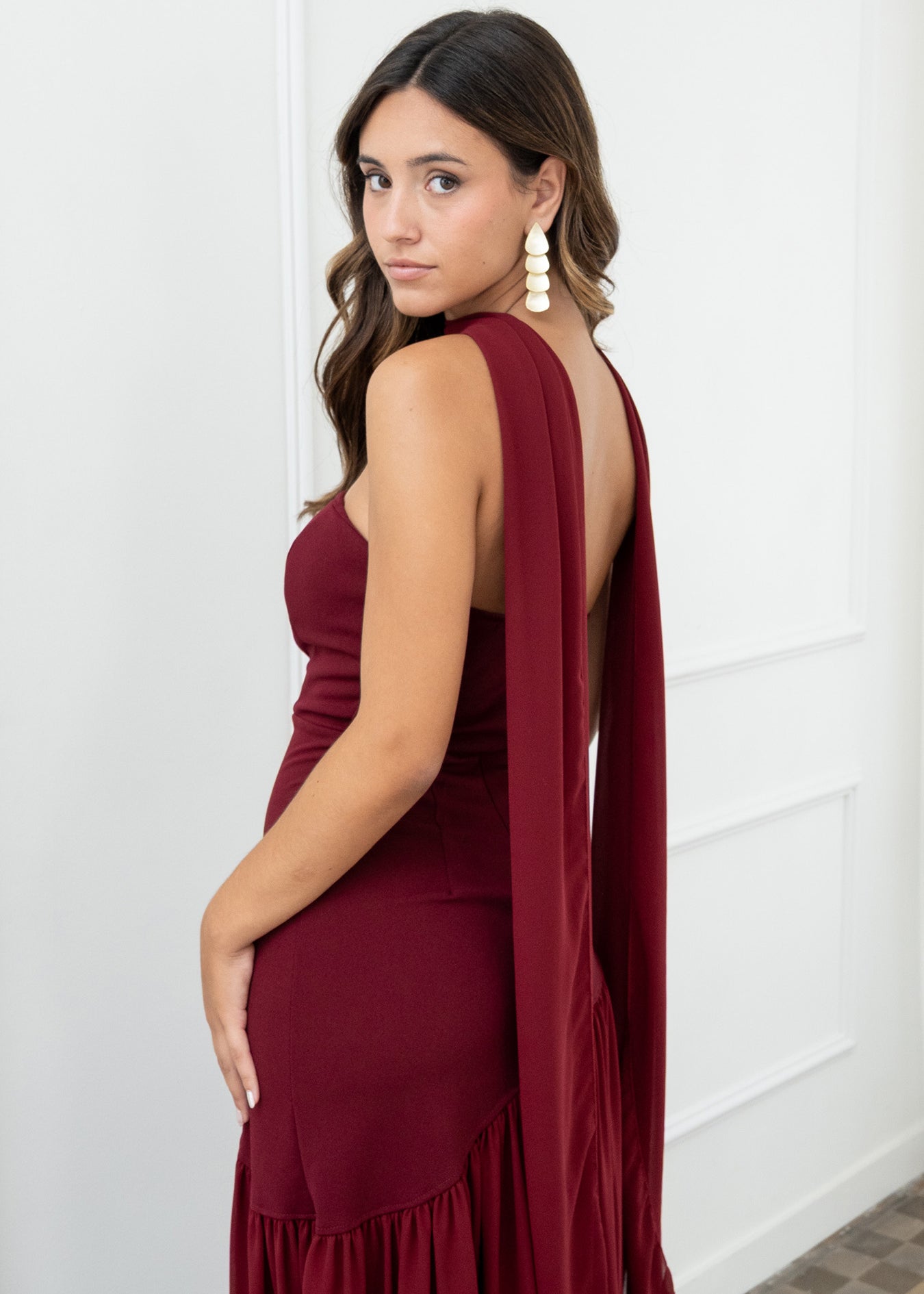 Modelo con vestido largo burdeos,  ideal para invitadas a bodas, fiestas de noche o eventos de gala. Escote palabra de honor que realza los hombros y el cuello, acompañado de un elegante fular integrado, aportando un toque sofisticado y original.