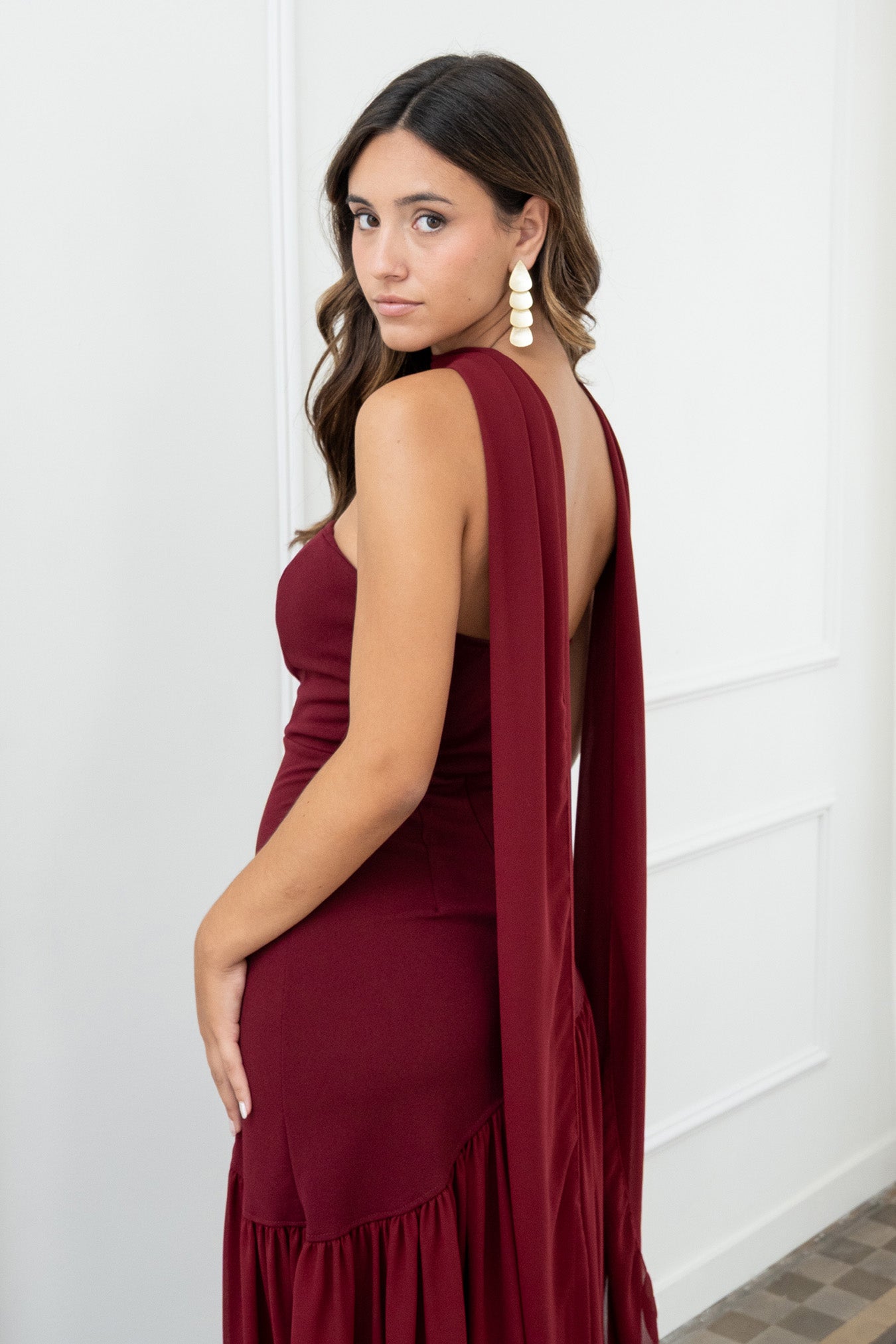Modelo con vestido largo burdeos,  ideal para invitadas a bodas, fiestas de noche o eventos de gala. Escote palabra de honor que realza los hombros y el cuello, acompañado de un elegante fular integrado, aportando un toque sofisticado y original.
