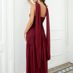 Modelo con vestido largo burdeos,  ideal para invitadas a bodas, fiestas de noche o eventos de gala. Escote palabra de honor que realza los hombros y el cuello, acompañado de un elegante fular integrado, aportando un toque sofisticado y original.
Confeccionado en un tejido fluido y ligero, el vestido presenta una falda amplia con vuelo y semitransparente  que añade movimiento y sensualidad al caminar.