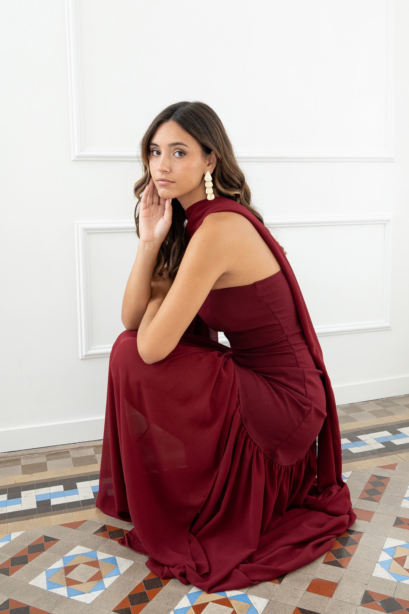 Modelo con vestido largo burdeos,  ideal para invitadas a bodas, fiestas de noche o eventos de gala. Escote palabra de honor que realza los hombros y el cuello, acompañado de un elegante fular integrado, aportando un toque sofisticado y original.
Confeccionado en un tejido fluido y ligero, el vestido presenta una falda amplia con vuelo y semitransparente  que añade movimiento y sensualidad al caminar.