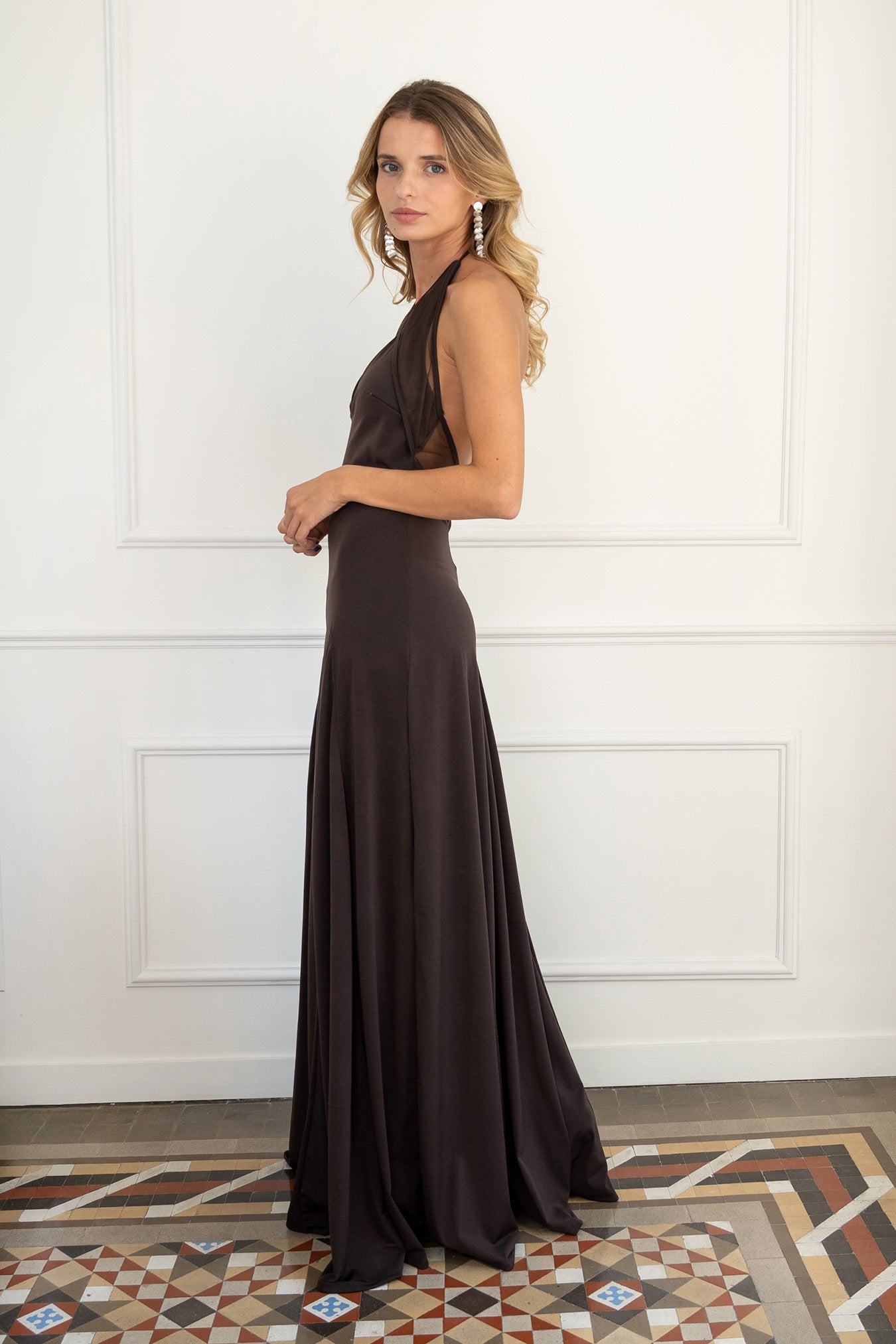 Modelo con vestido marrón chocolate, confeccionado en un tejido de licra con cuerpo que se adapta a la figura. Su escote halter y las delicadas piezas de tul en el cuello y la espalda abierta le dan un toque romántico y sutil.
La falda midi, con godetes que le aportan un movimiento fluido y ligero.