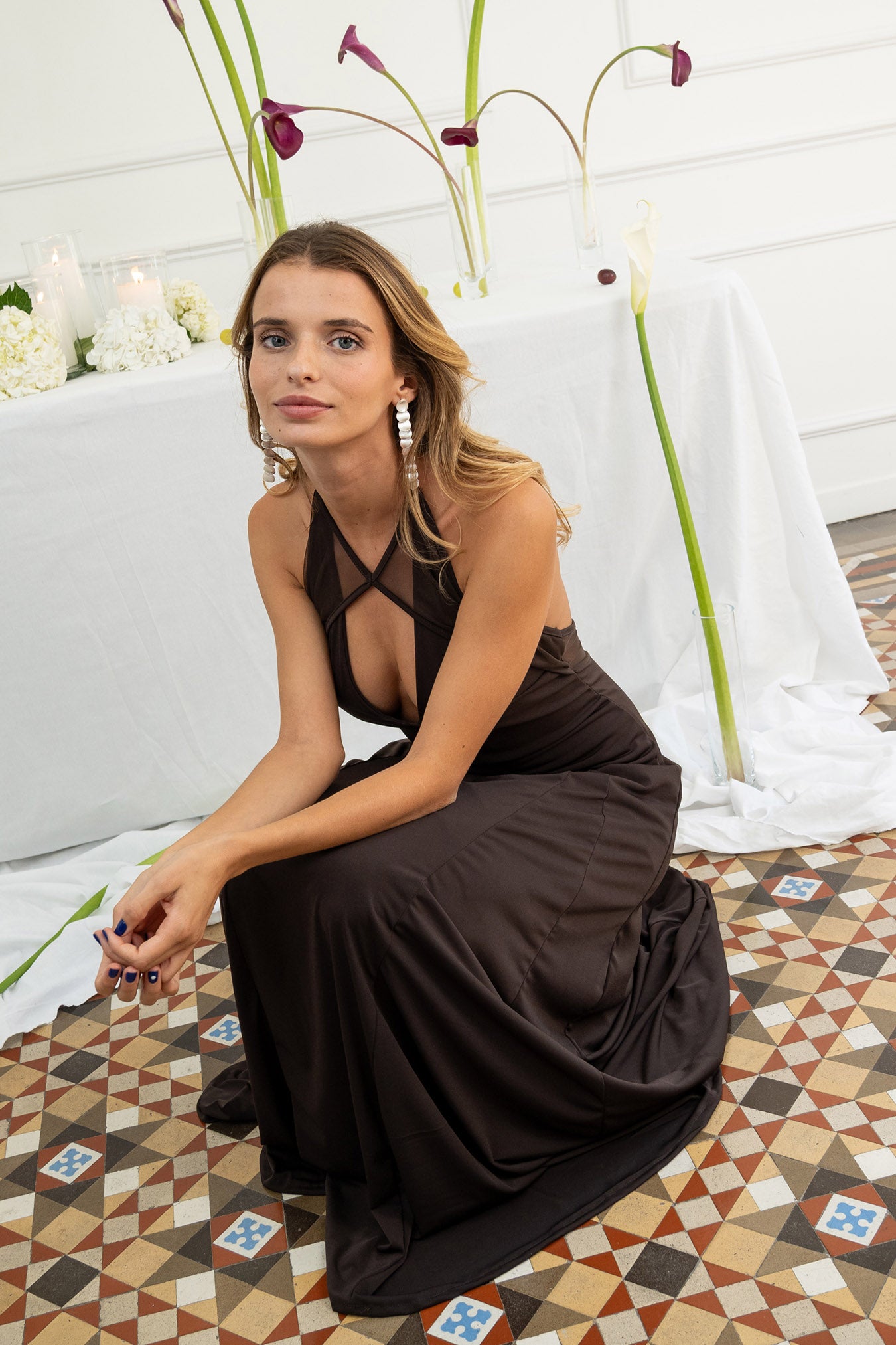 Modelo con vestido marrón chocolate, confeccionado en un tejido de licra con cuerpo que se adapta a la figura. Su escote halter y las delicadas piezas de tul en el cuello y la espalda abierta le dan un toque romántico y sutil.
La falda midi, con godetes que le aportan un movimiento fluido y ligero.