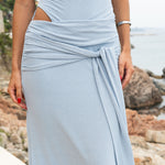 Mujer con vestido largo azul claro y corte elegante, posando de pie en la costa con vistas al mar y rocas de fondo.