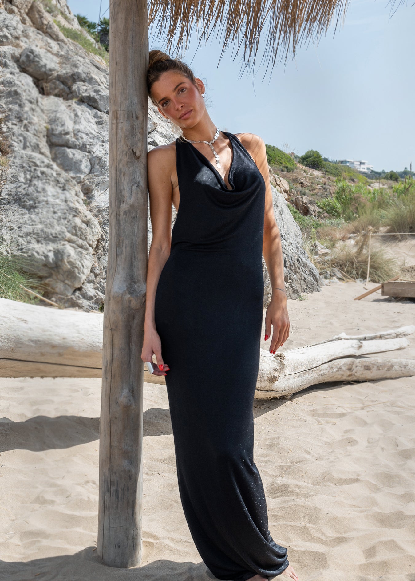 Vestido largo negro de lurex con escote drapeado y collar plateado en la playa