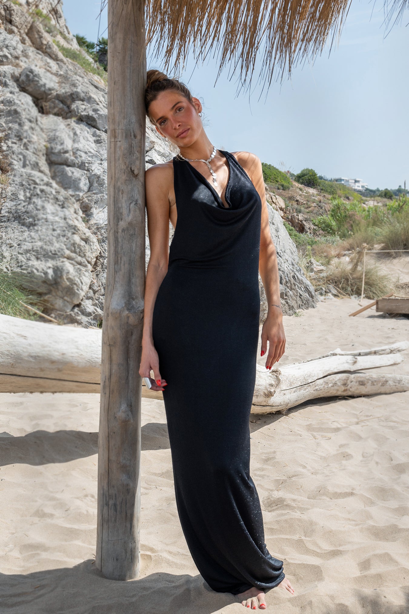 Vestido largo negro de lurex con escote drapeado y collar plateado en la playa