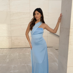 Modelo con vestido largo azul celeste de escote asimétrico cruzado y silueta fluida, posando frente a una pared de piedra clara.