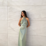 Modelo con vestido largo color salvia de escote halter cruzado y silueta fluida, posando frente a una pared de piedra clara.