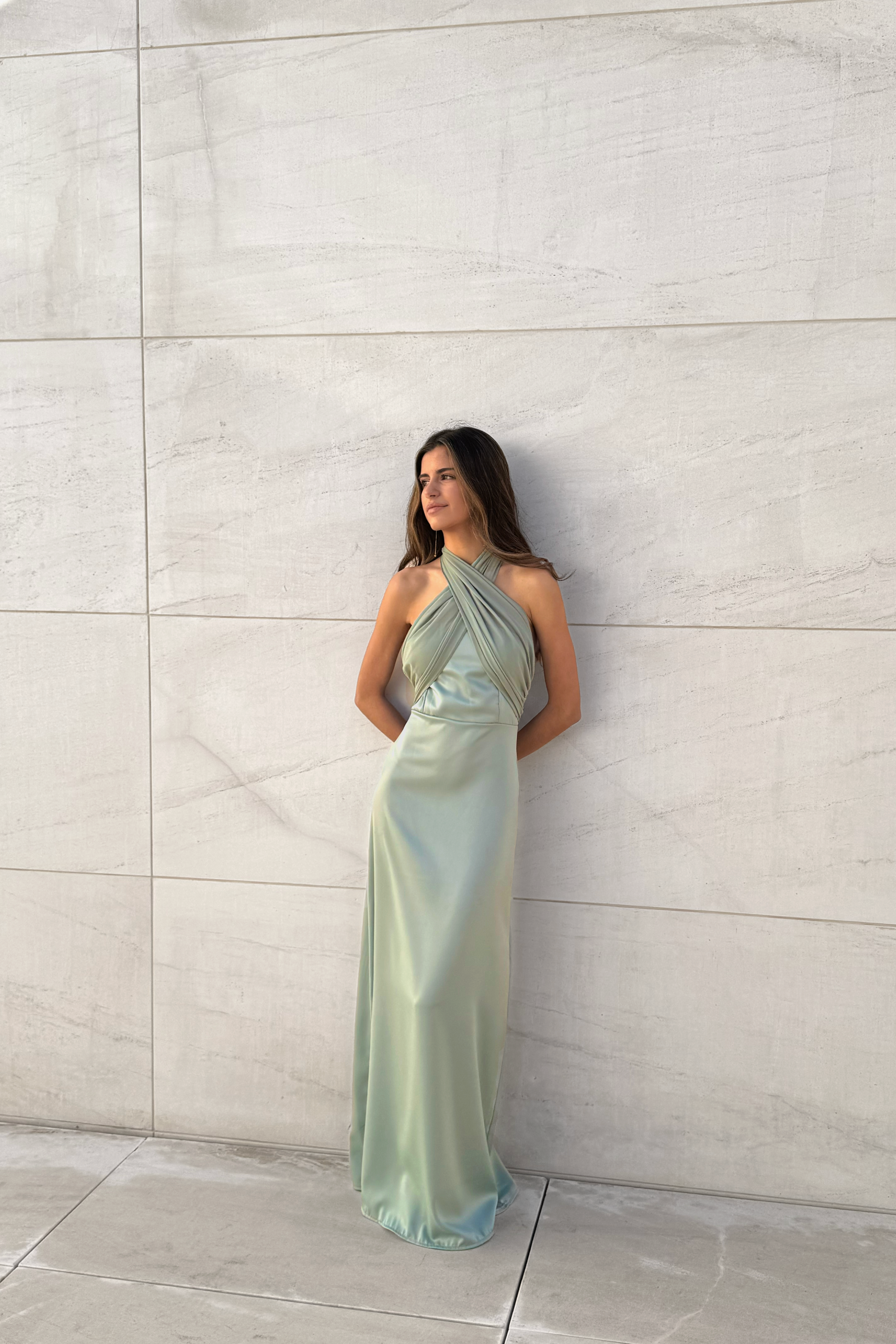Modelo con vestido largo color salvia de escote halter cruzado y silueta fluida, posando frente a una pared de piedra clara.