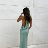 Modelo con vestido largo color salvia de escote halter y espalda descubierta, silueta fluida, vista posterior frente a una pared de piedra clara.