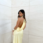 Modelo con vestido largo amarillo de escote halter y espalda descubierta, de tejido fluido, posando frente a una pared de piedra clara.